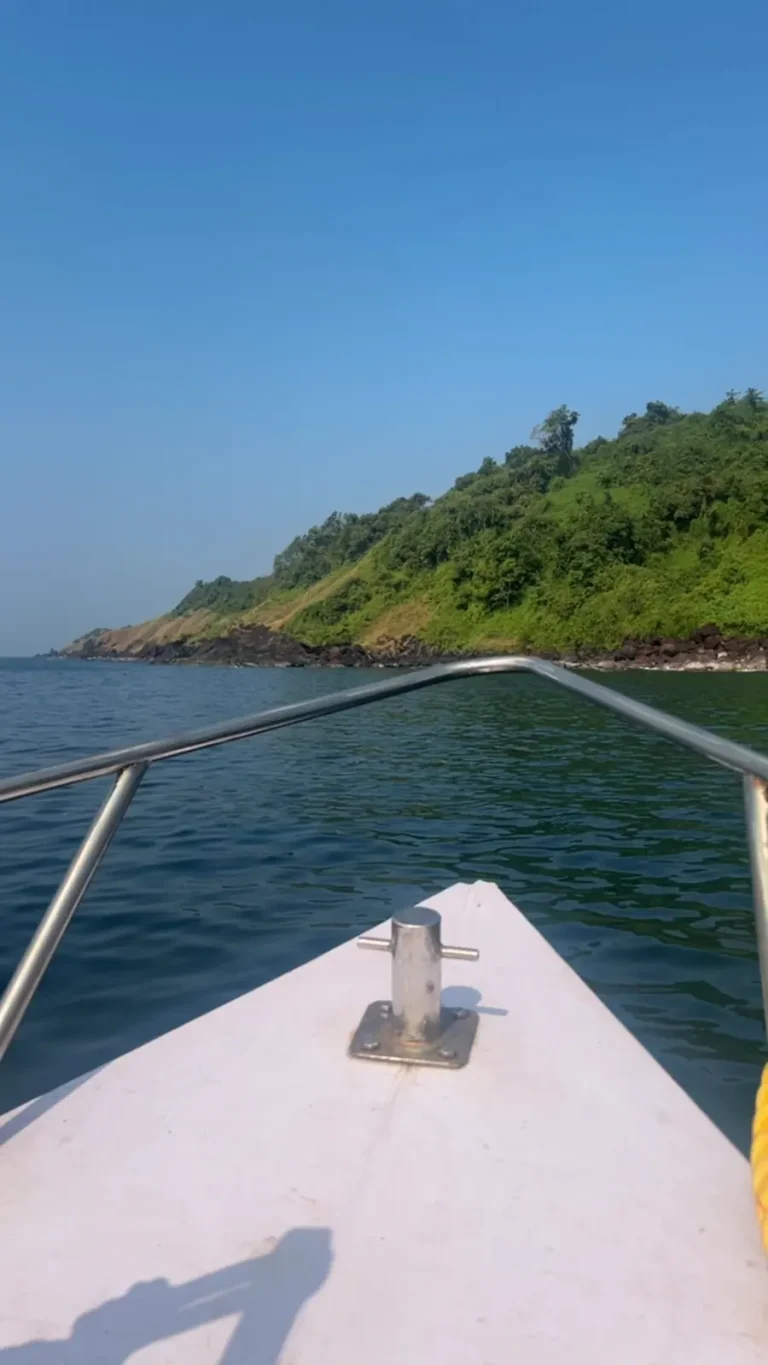 View over the white bow rail of a dive boat heading across calm water toward a green headland