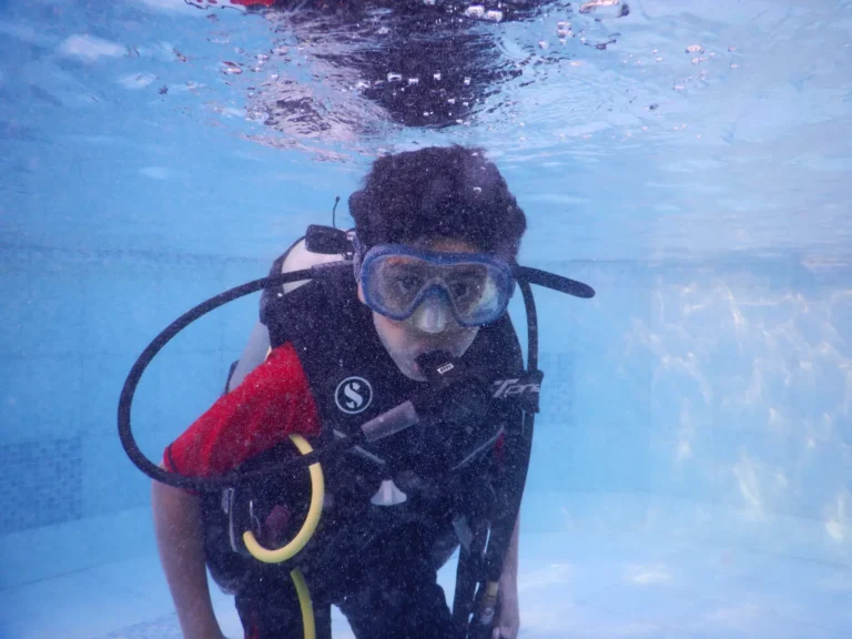 Boy in a mask, red rash guard and BCD kneels on the bottom of a pool during training