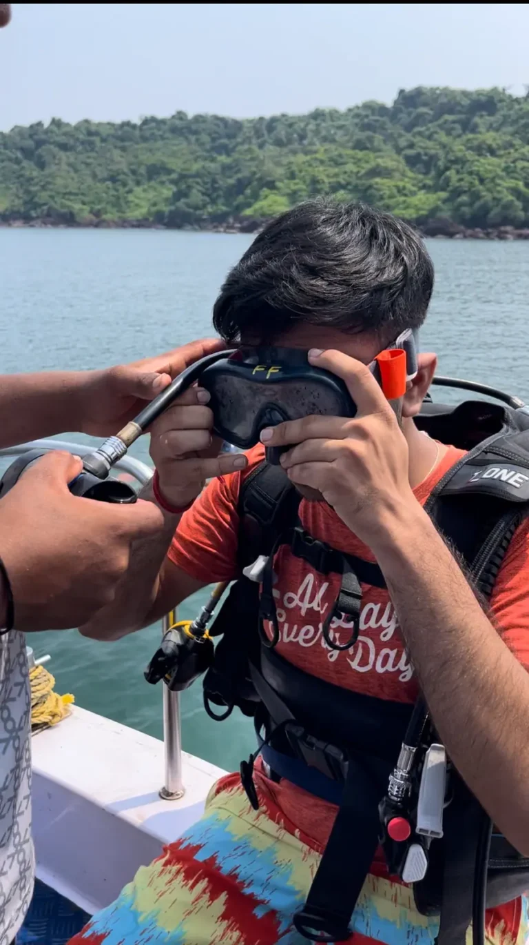 Boy in a red T-shirt and BCD pulls on his mask aboard a boat, with a green island behind