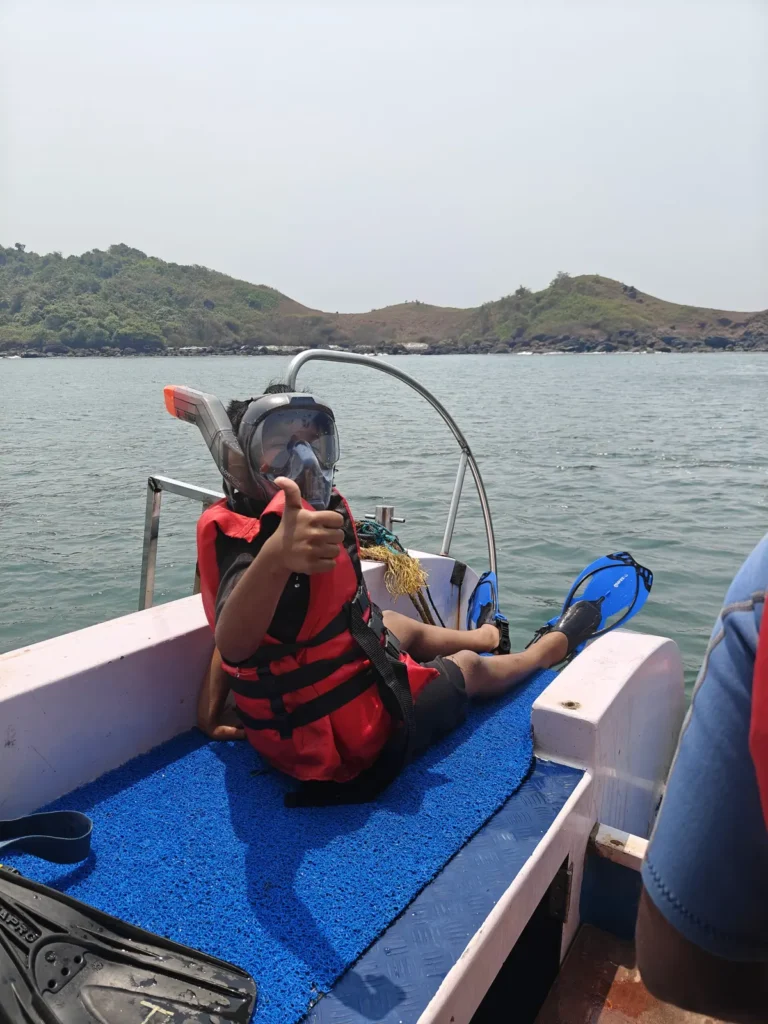 A child in a red life jacket and snorkel mask gives a thumbs up from a boat seat, with hills behind