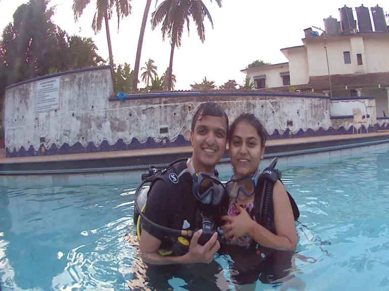 Smiling couple in BCDs hold their masks while standing in the pool, palm trees behind