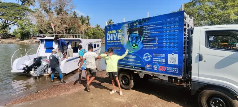 Crew unload gear from a truck with a Learn to Dive banner onto a boat moored at a riverside jetty