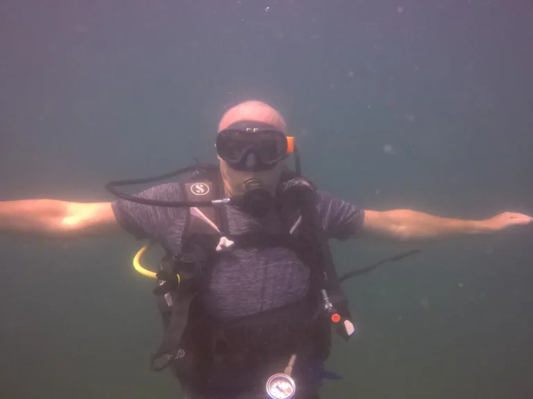 Diver in a dark mask holds his arms straight out to the sides while hovering in hazy open water