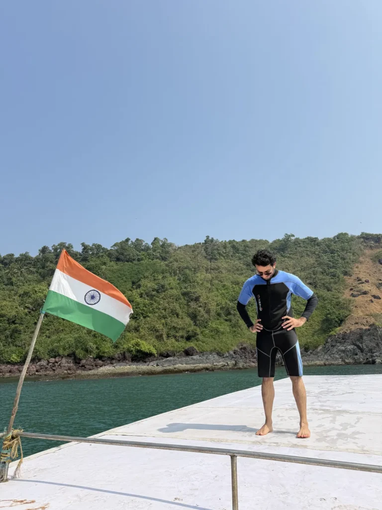 Man in a shorty wetsuit stands on the boat bow beside an Indian flag, with a green hillside behind