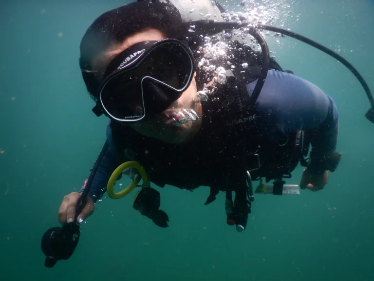 Diver in a black mask swims toward the camera with a stream of bubbles rising past his face