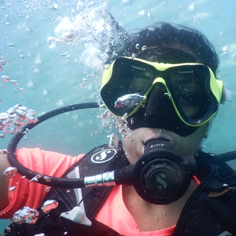 Diver in a yellow-rimmed mask and pink top surrounded by bubbles, with small fish in the background
