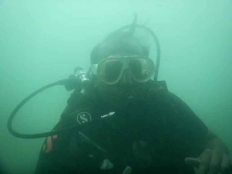 Close view of a diver in a clear mask and black BCD pointing toward the camera in green water
