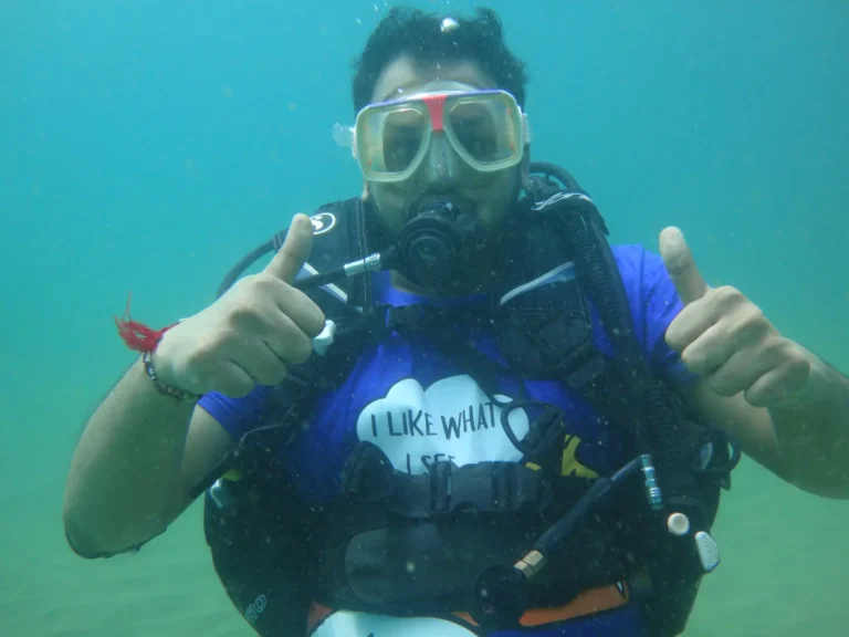 Diver in a blue T-shirt gives two thumbs up while hovering over a sandy sea floor