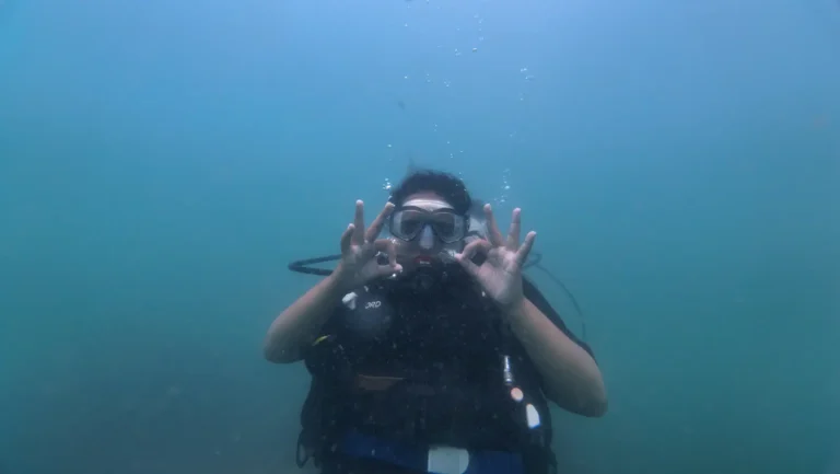 A diver hovering in blue water makes circles with her fingers around her mask