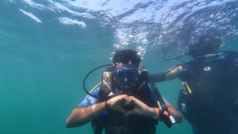 Diver in a blue mask makes heart hands under the surface while his instructor stays close beside him