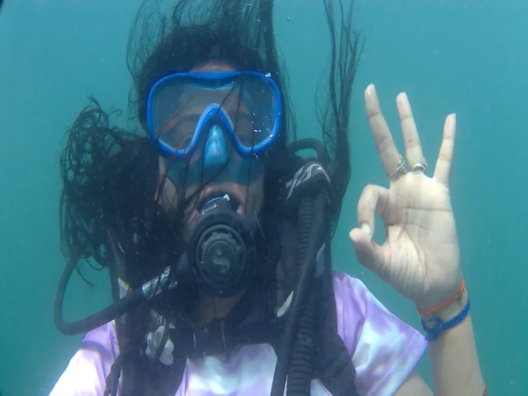 A woman diver with hair floating around her blue mask gives a clear OK sign in open water