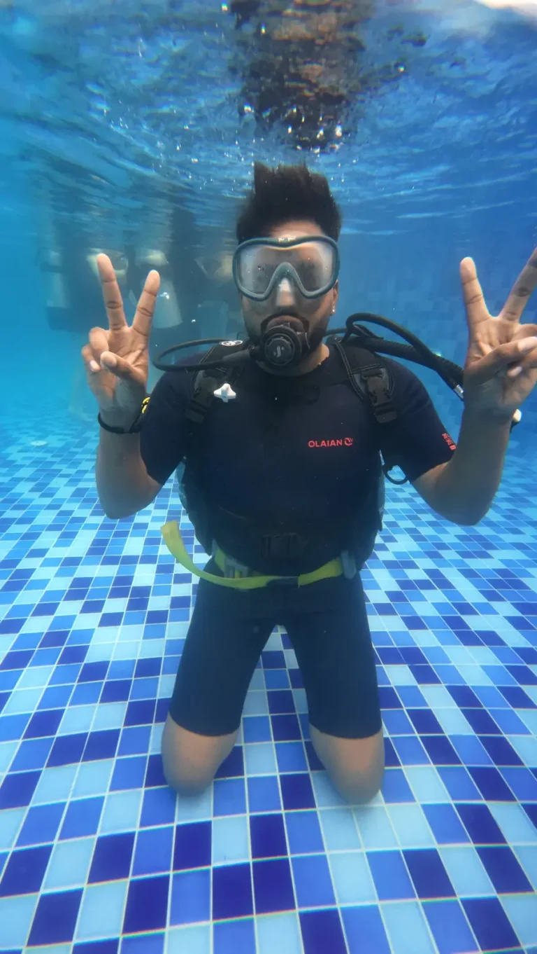 Diver in a black shorty and mask kneels on a tiled pool floor, flashing peace signs with both hands