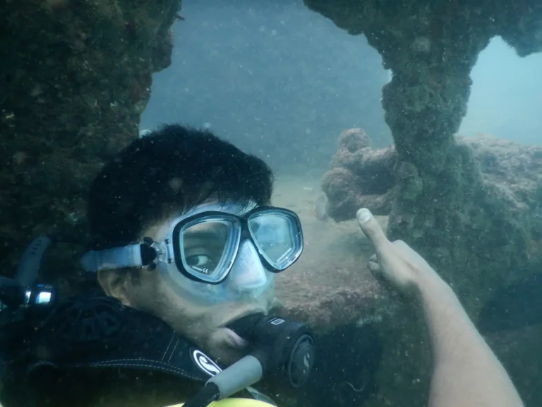 A young diver in a clear mask points back toward encrusted wreck beams behind him