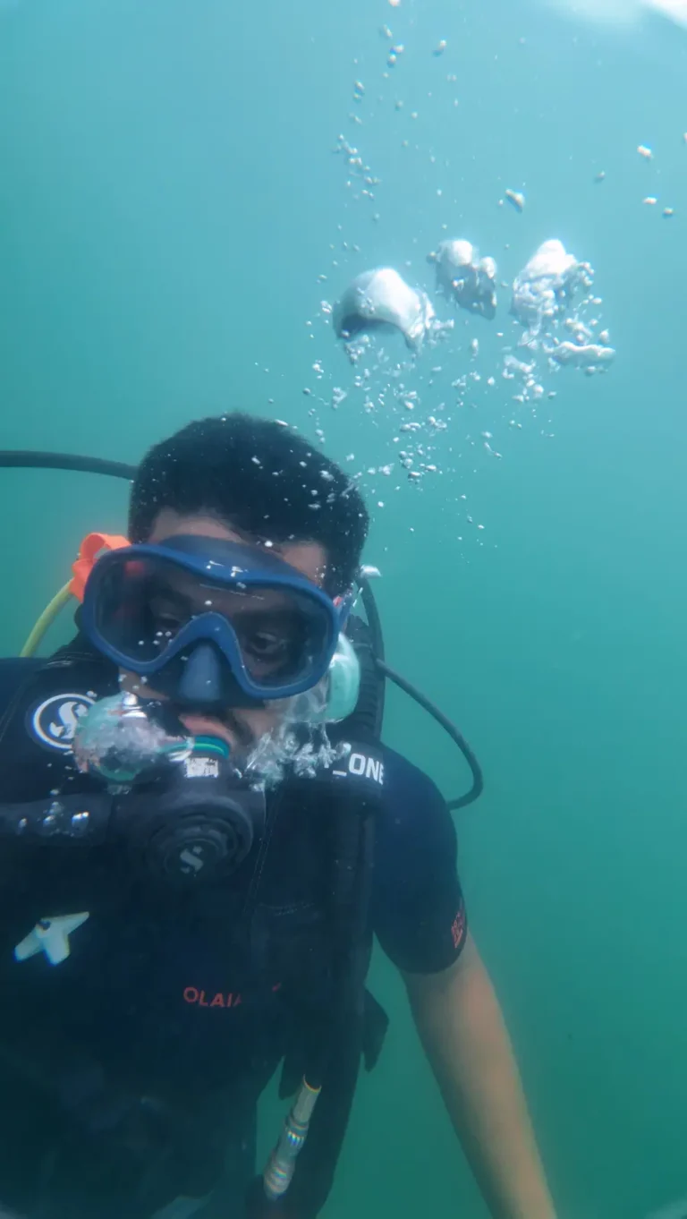 Diver in a blue mask and black T-shirt hovers in green-blue water as bubbles rise past him