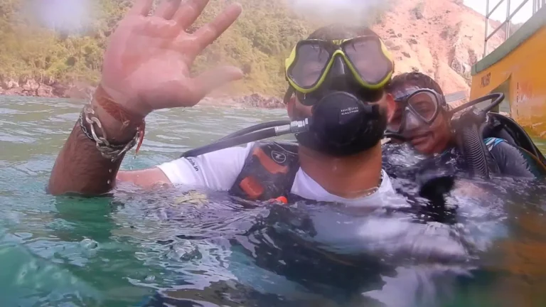 Diver in a yellow mask waves at the camera at the surface, with a buddy and the boat behind