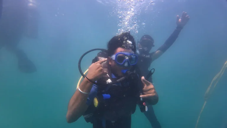 Diver touches his blue mask while a buddy behind him waves at the camera in blue water