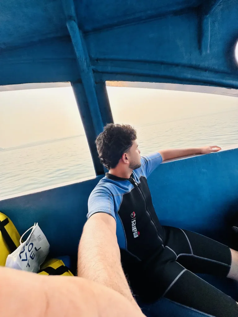 A diver in a shorty wetsuit sits by an open boat window, looking out over the sea in soft morning light