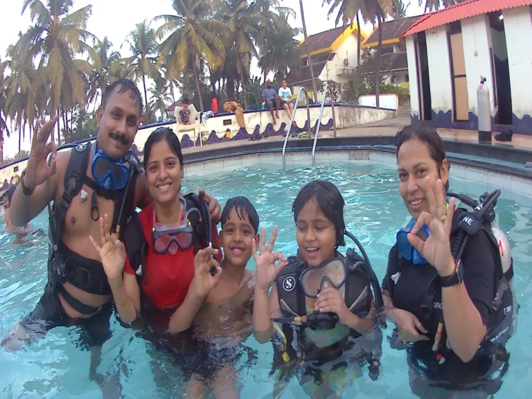 Family of five in BCDs and masks give OK signs together in the resort pool, palms behind