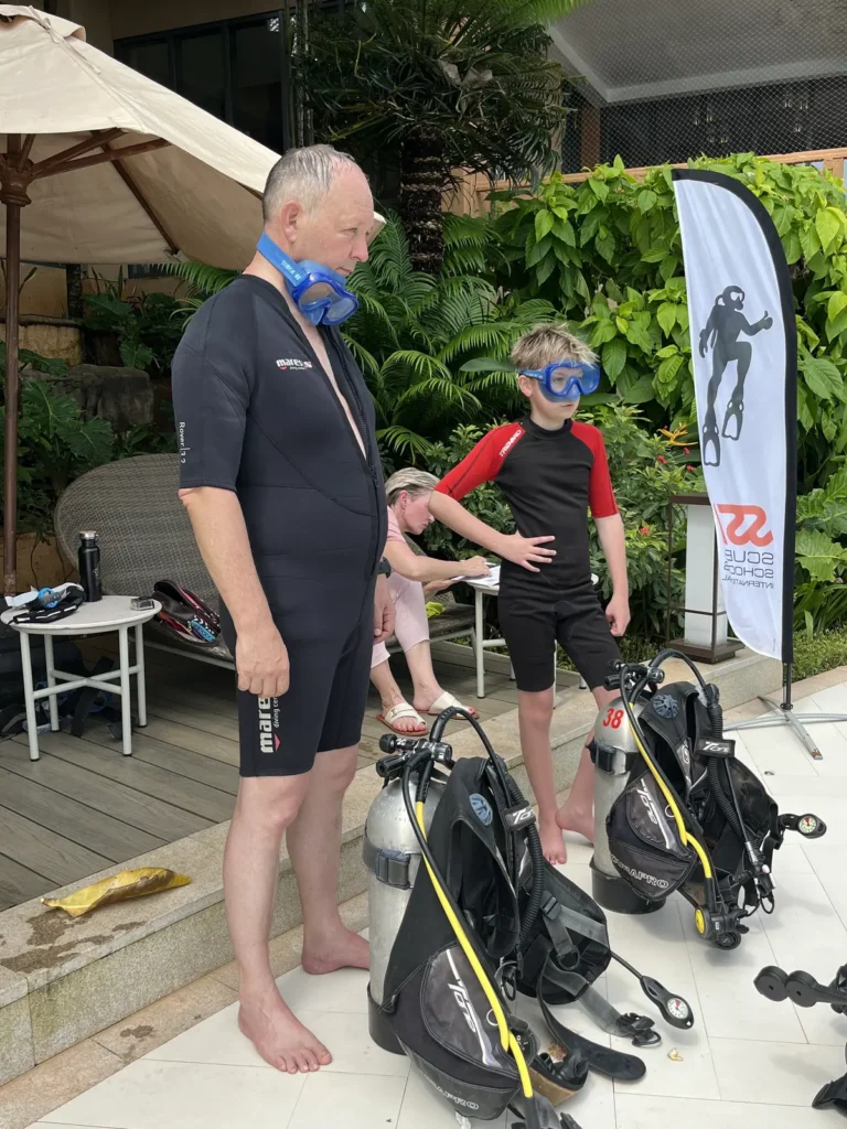 Man and boy in wetsuits stand beside two assembled scuba kits on a poolside deck