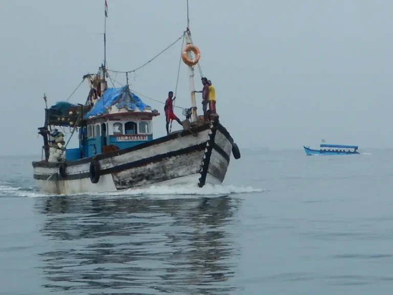 A wooden fishing trawler with crew on deck moves across flat grey water, a smaller boat in the distance