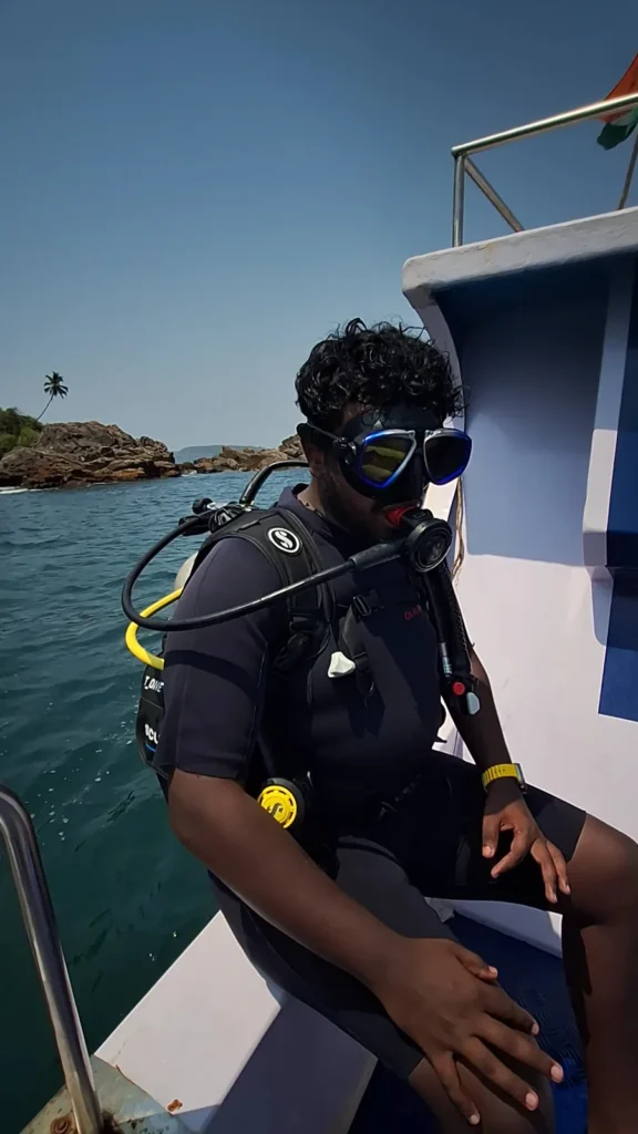 Diver in full scuba kit and mask seated on the edge of a boat with a rocky island behind