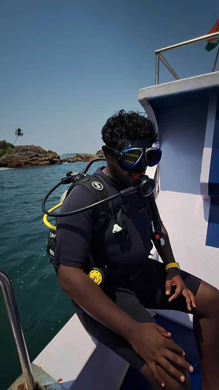 Diver in full scuba kit and mask seated on the edge of a boat with a rocky island behind