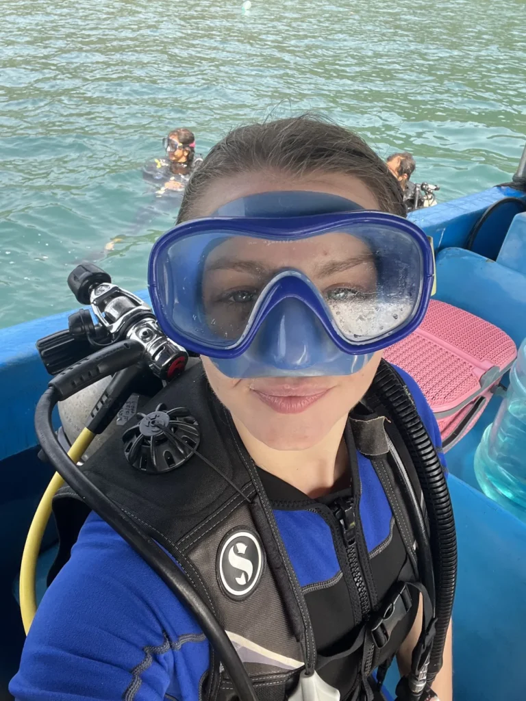 Woman in a blue mask and Scubapro BCD taking a selfie on the boat with divers in the water behind