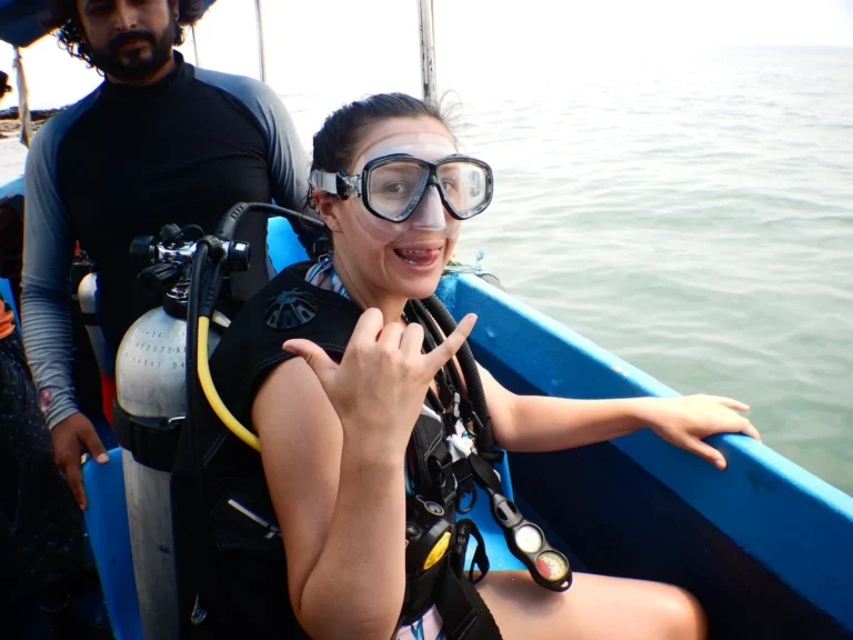 Woman in mask and scuba gear on a dive boat making a shaka sign, instructor behind