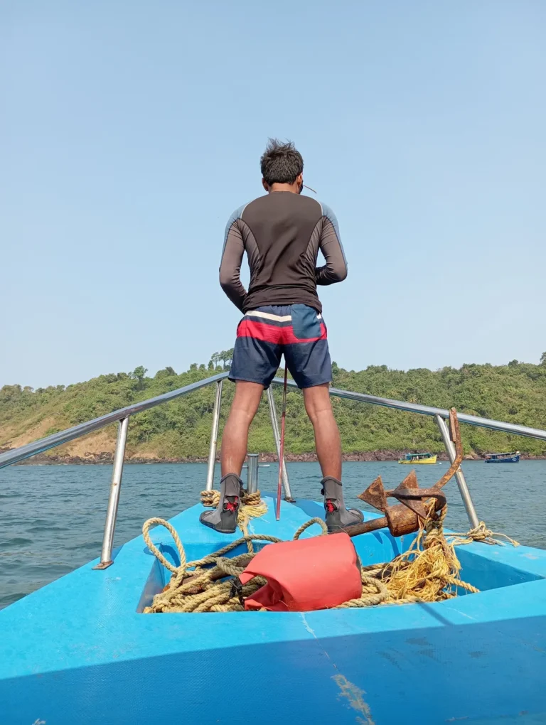 Crew member on a blue boat's bow with anchor and rope, approaching a green island