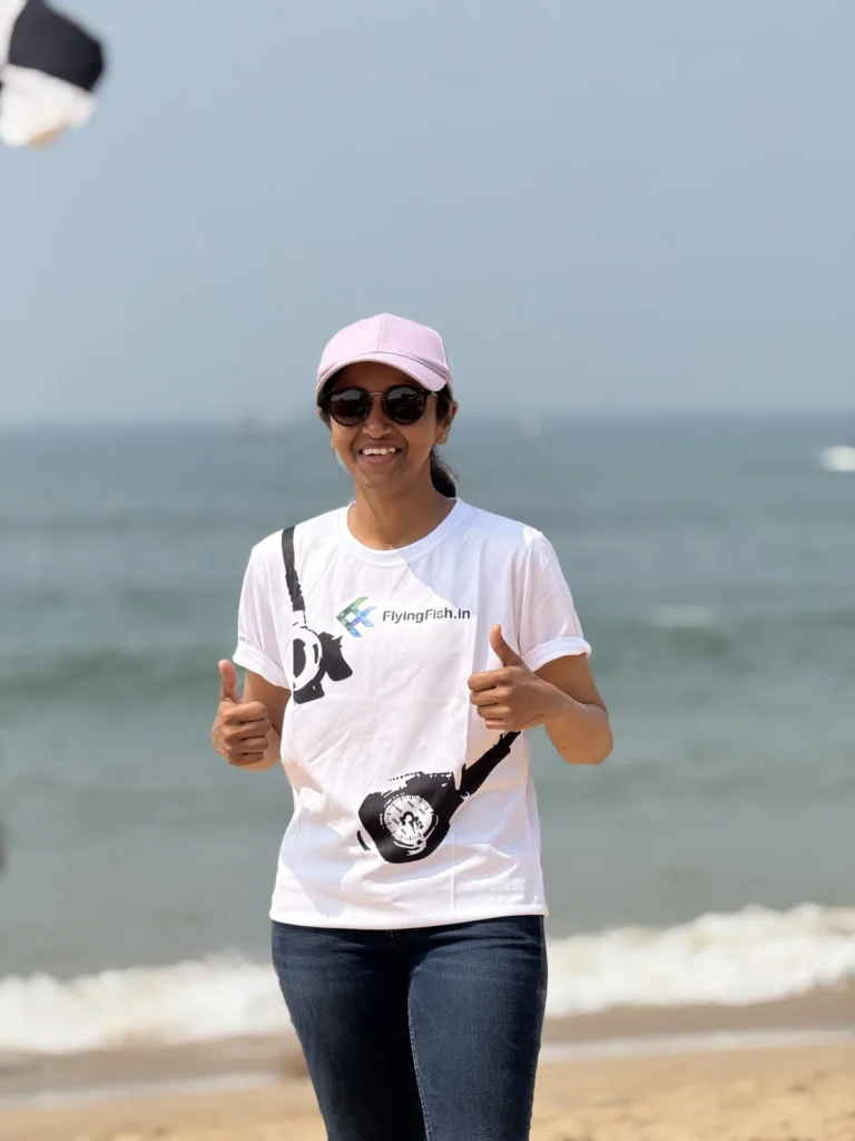 Smiling woman in a FlyingFish.in t-shirt and cap giving a thumbs up on the beach