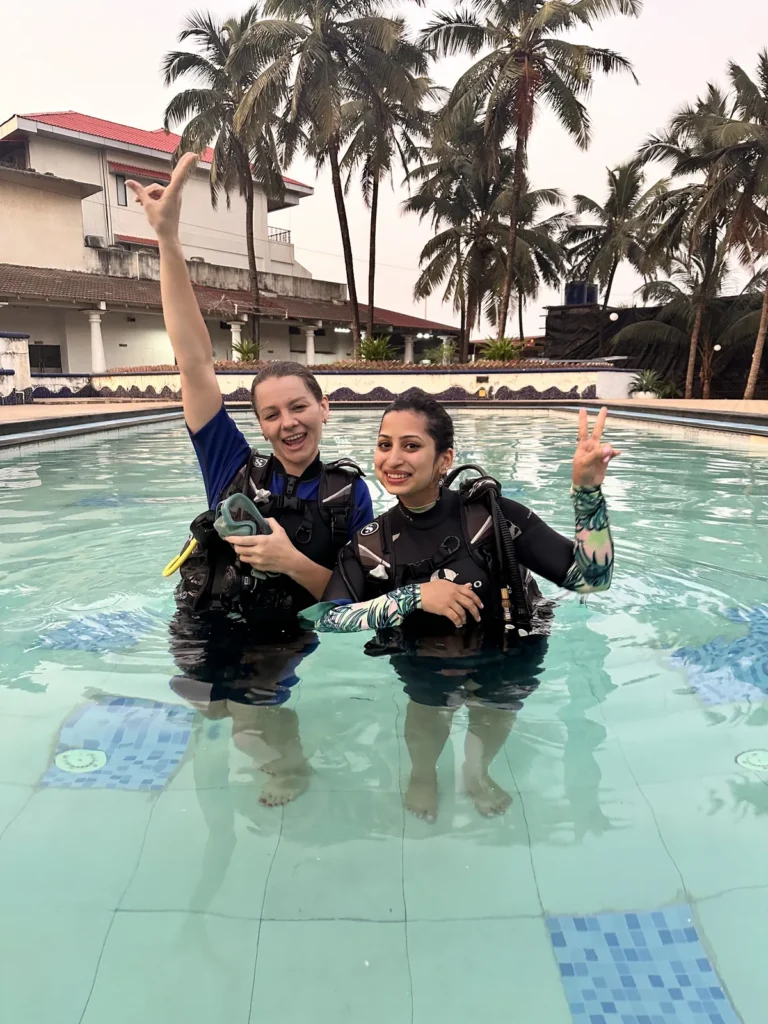 Two smiling women in BCDs standing waist-deep in a resort pool with palm trees behind