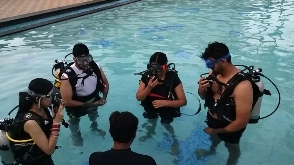 Instructor briefs four students in scuba kit standing in a pool