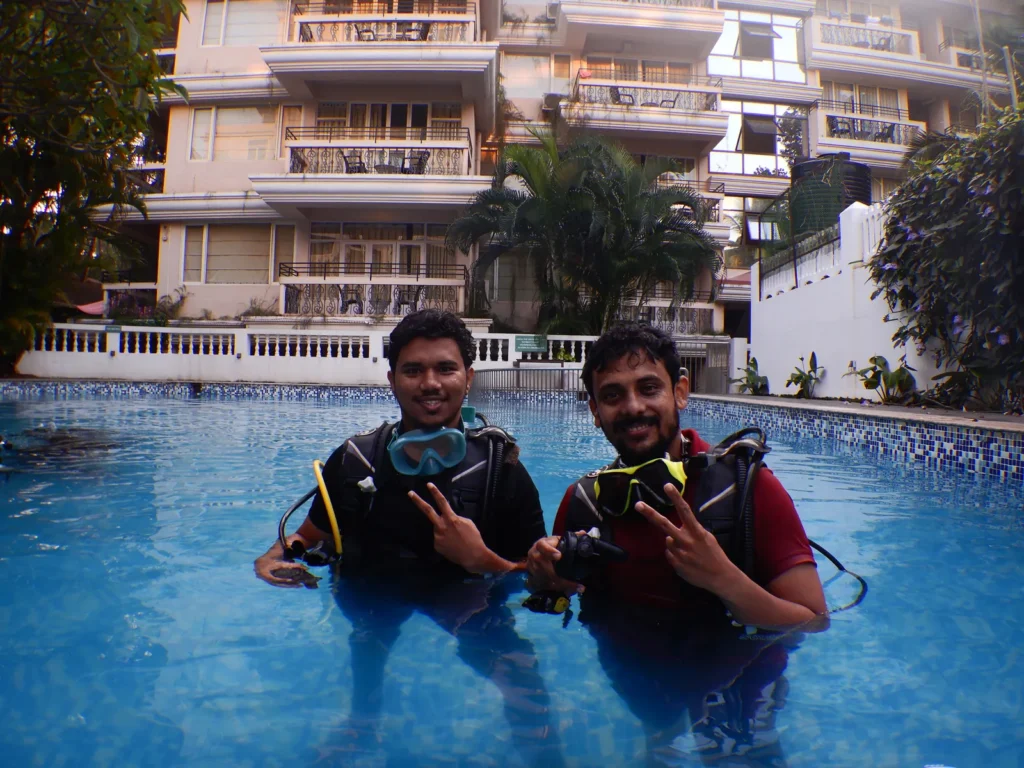 Two male guests in scuba gear giving peace signs in a pool in front of an apartment building