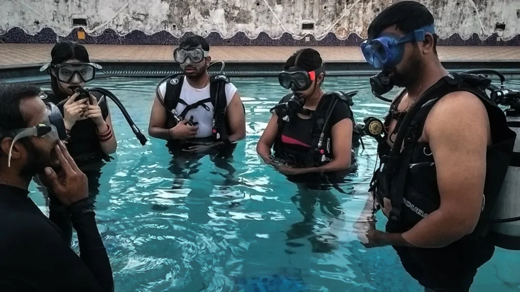 Five students in scuba gear standing in a pool during a briefing, with an instructor in the foreground
