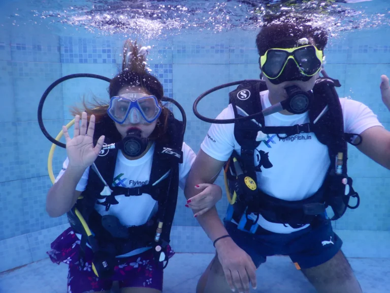 Two divers in FlyingFish.in t-shirts pose underwater in a pool, one waving