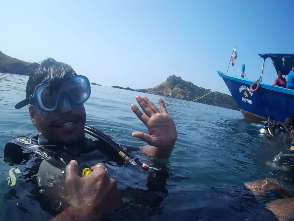 Smiling diver waves at the sea surface with a dive boat and island behind