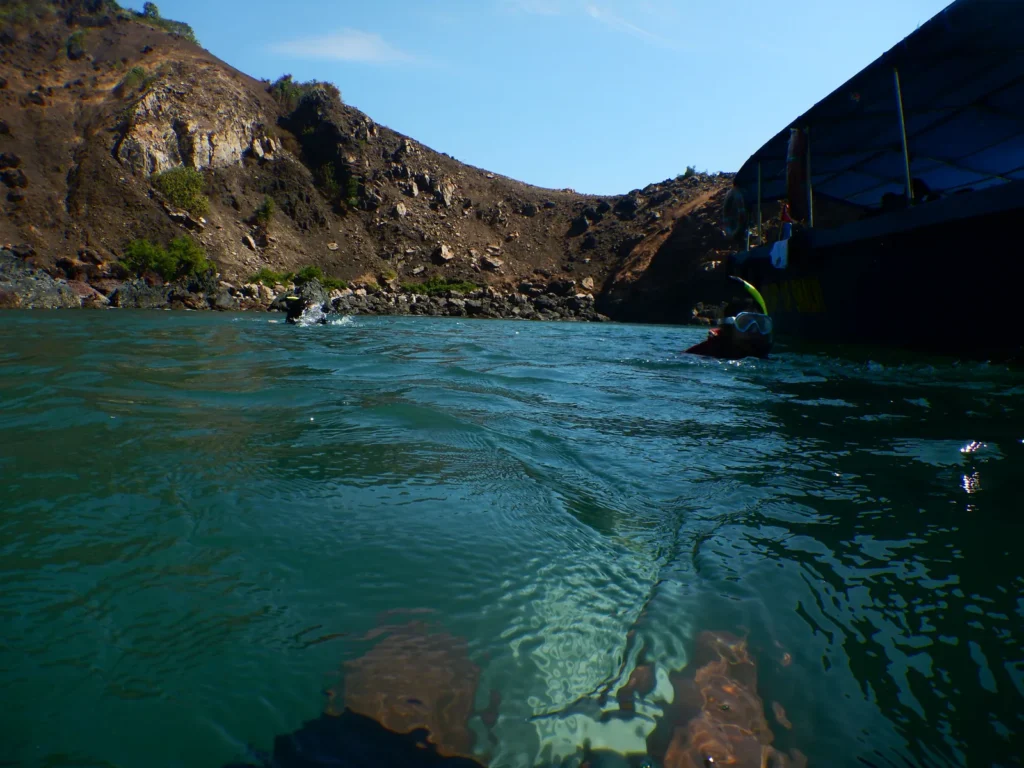 Water-level view of divers swimming near a rocky cove and the dive boat