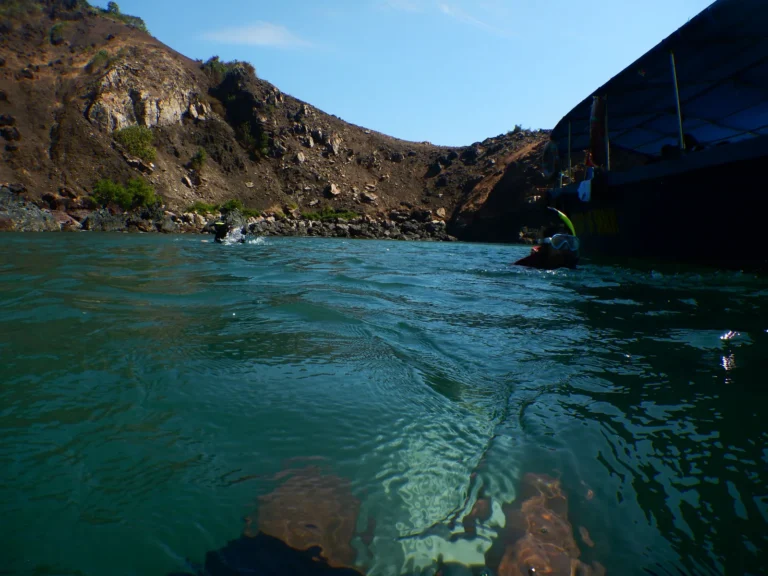 Water-level view of divers swimming near a rocky cove and the dive boat