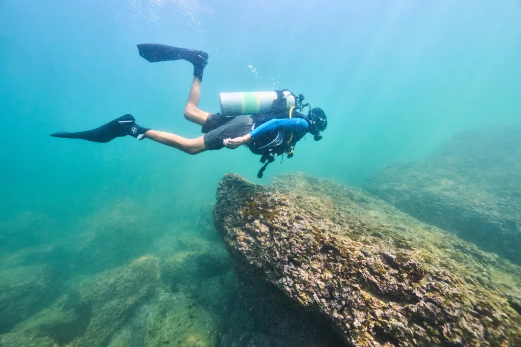 Diver in full gear finning over a rocky reef in clear blue-green water