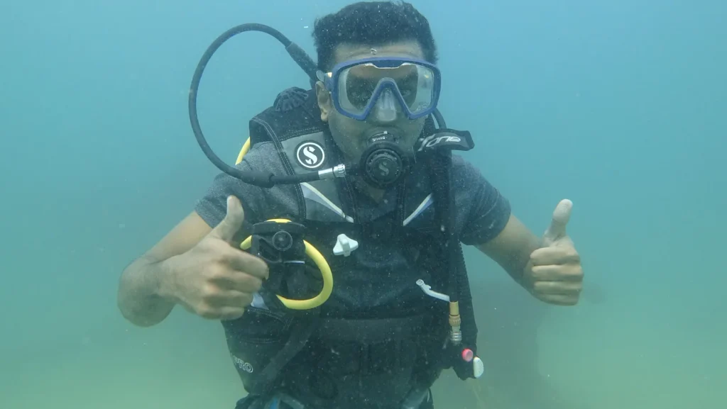 Diver in a Scubapro BCD gives two thumbs up underwater in turquoise water