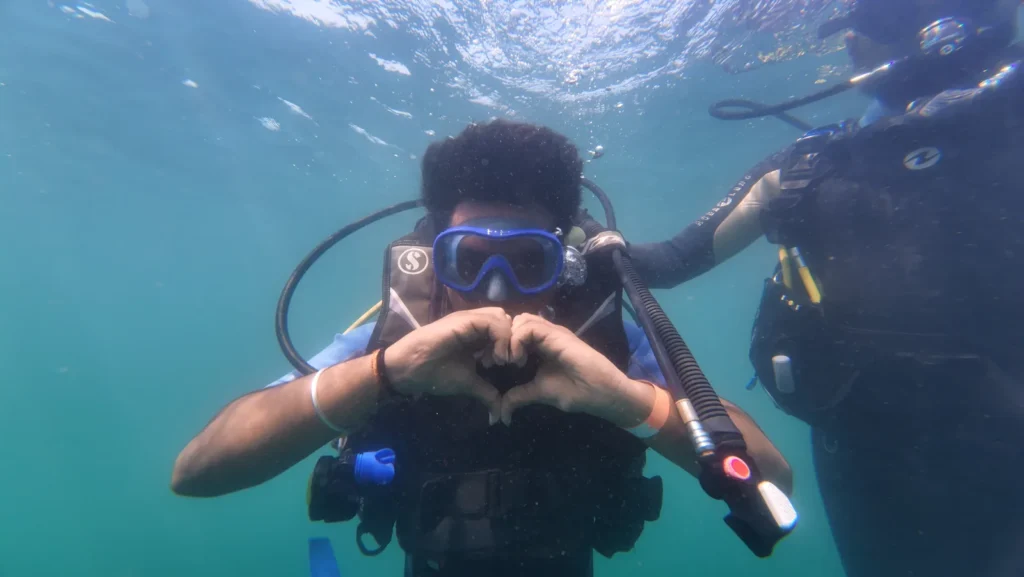 Diver making a heart sign with his hands underwater while an instructor holds him from the side