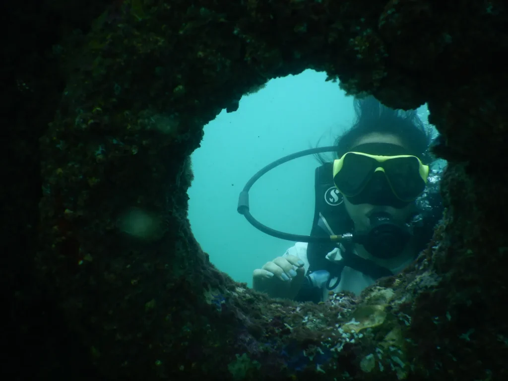 Diver in a yellow mask seen through a round encrusted porthole of a shipwreck