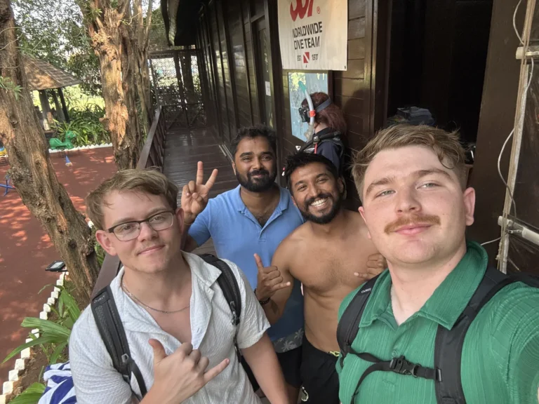 Four smiling young men take a selfie on the wooden veranda of the dive centre, flashing hand signs