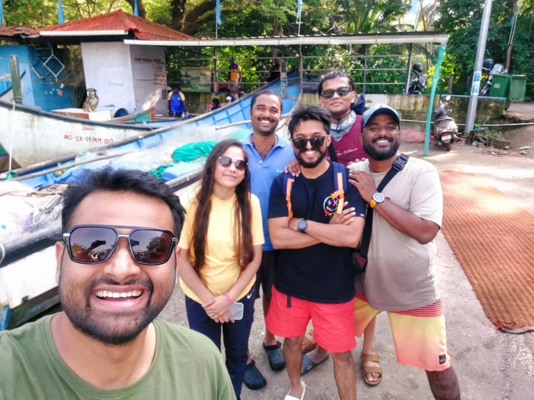 Group of six smiling friends take a selfie beside fishing boats at the jetty before heading out