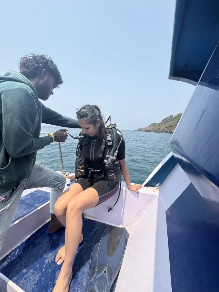 Crew member checks the gear of a woman in a BCD seated on the boat edge near a green headland