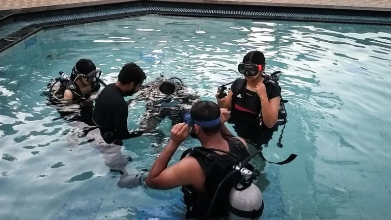 Instructor checks a student's kit while three other students in masks and BCDs wait in the pool