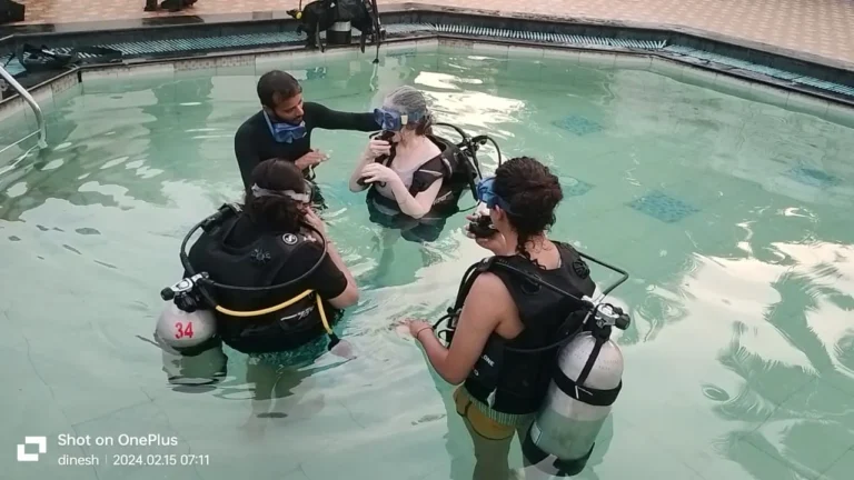 An instructor adjusts a student's mask in a shallow pool while two other students in full gear wait