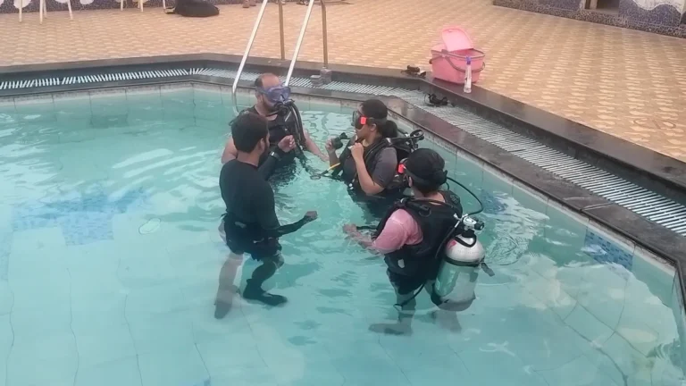 Instructor stands waist deep in a resort pool with three students in scuba kits, going over the skills