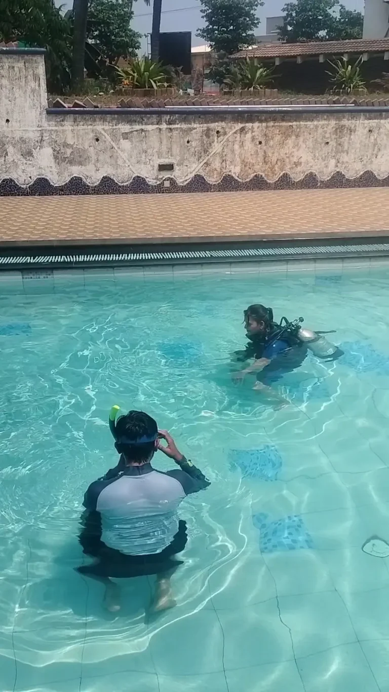 Instructor crouches in the shallow end watching a student in scuba gear swim across the pool