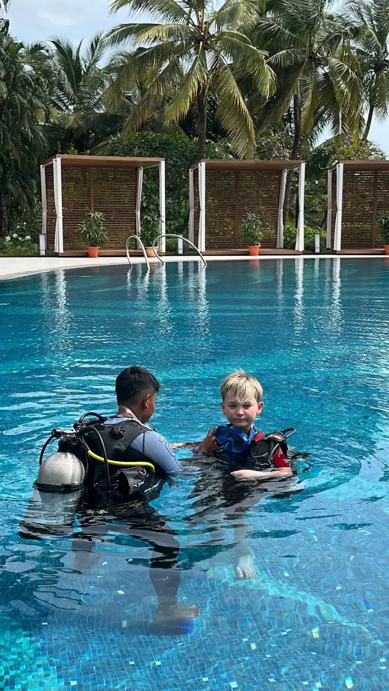 An instructor stands in a palm-lined pool beside a young boy wearing a small scuba jacket and tank